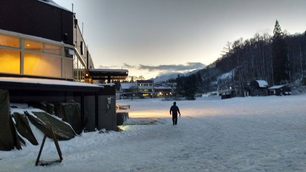 A man walks across an expanse of snow past a large building on his left