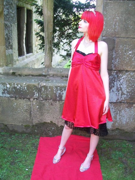 A girl with bright red hair wearing a bright red dress, standing in front of a window in a ruined building